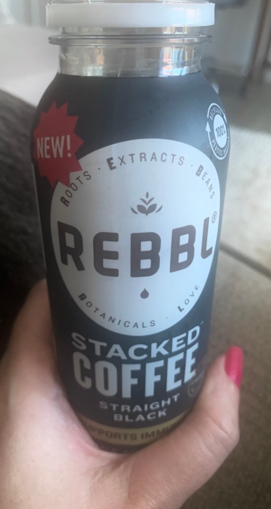 A hand with pink nail polish holding a REBBL Stacked Coffee Straight Black beverage.